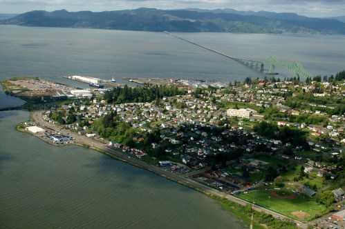 Aerial view of a coastal town with a bridge, waterfront, and green hills in the background.