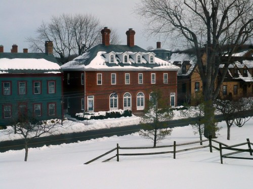 A snowy landscape featuring a brick house with multiple chimneys, surrounded by trees and other colorful homes.