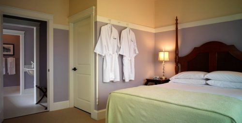 Cozy bedroom with a bed, lamp, and hanging bathrobes; bathroom visible through an open door. Soft purple walls.
