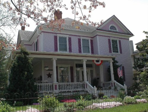 A charming two-story house with a porch, purple shutters, and a well-kept garden, featuring American flags and decorative elements.
