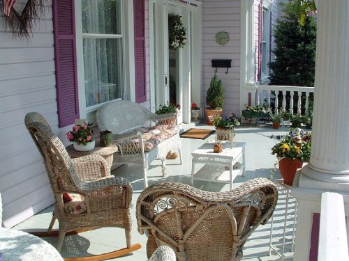 A cozy porch with wicker furniture, potted plants, and a welcoming atmosphere, featuring pink walls and white railings.