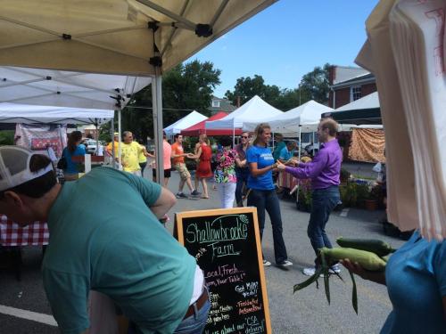 A bustling farmers market with colorful tents, people shopping, and a chalkboard sign for a local farm.