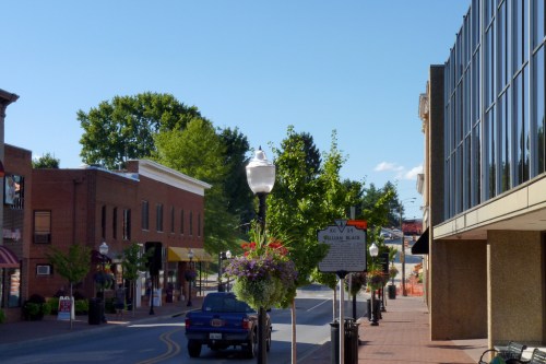 A quiet street scene with shops, trees, and a blue sky, featuring hanging flower baskets and a parked blue truck.