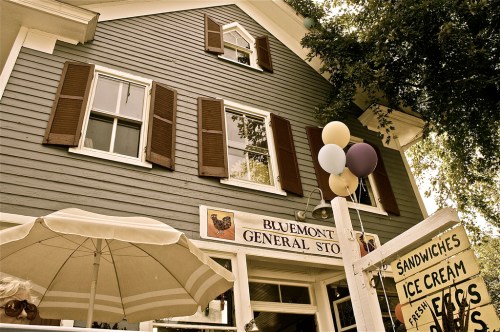 A charming general store with a blue exterior, shutters, and a sign, featuring balloons and an umbrella in the foreground.