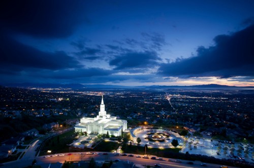 Aerial view of a lit temple at dusk, surrounded by a cityscape and mountains under a cloudy sky.