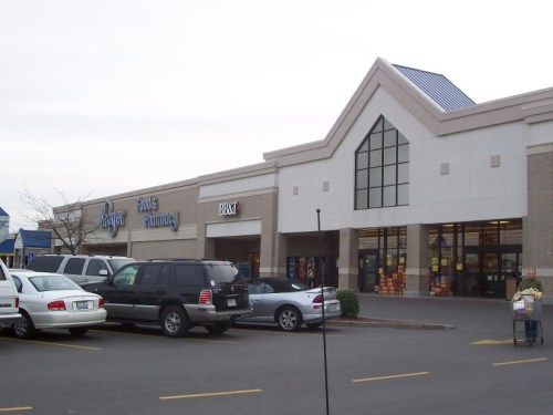 A storefront featuring a pharmacy and a grocery store, with parked cars in the foreground and a cloudy sky above.