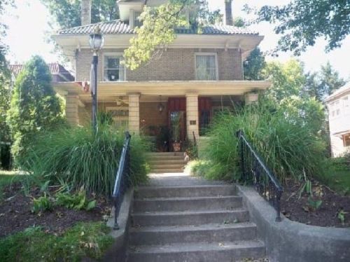A charming two-story brick house with a front porch, surrounded by greenery and a landscaped yard.