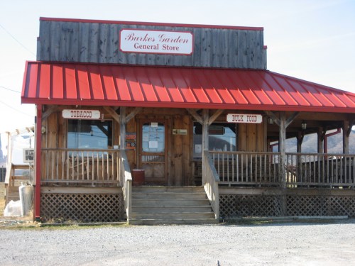 A rustic general store with a red roof and wooden exterior, featuring a porch and signs for tobacco and bulk food.