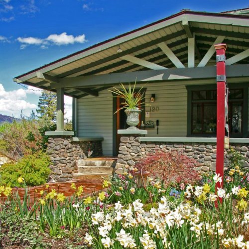 A charming house with a stone facade, surrounded by colorful flowers and greenery, under a bright blue sky.