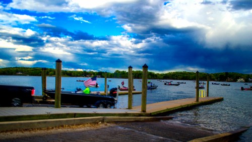 A scenic view of a lake with boats, a cloudy sky, and a dock lined with wooden posts.