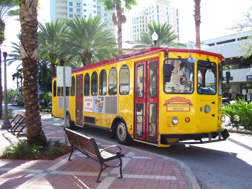 A bright yellow trolley bus parked on a palm-lined street with benches and buildings in the background.
