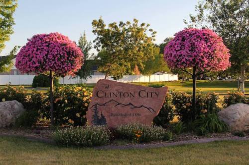 A stone sign for Clinton City, established 1896, surrounded by vibrant pink flowering trees and green landscaping.