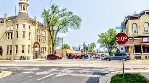A street view featuring a stop sign, historic buildings, and people gathered near a town square on a sunny day.