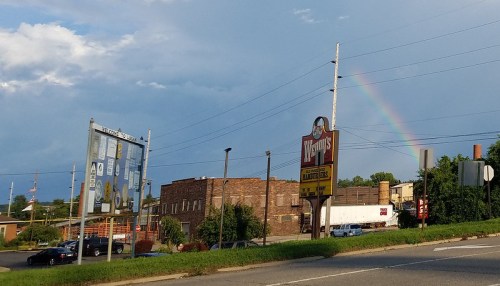 A Wendy's sign stands beside a road with a rainbow visible in the background against a cloudy sky.
