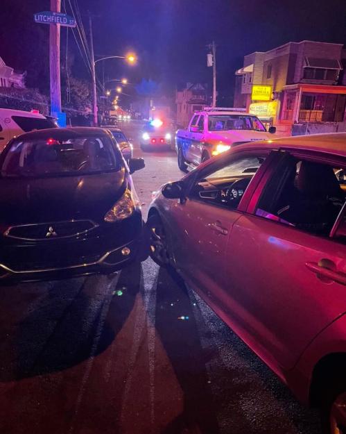 Two cars are parked closely together on a dimly lit street, with police lights flashing in the background.