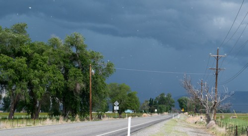 A rural road lined with trees under a dark, stormy sky, with power lines and distant mountains visible.