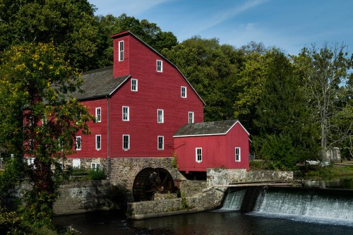 A red historic mill beside a river, featuring a waterwheel and surrounded by lush greenery under a clear blue sky.