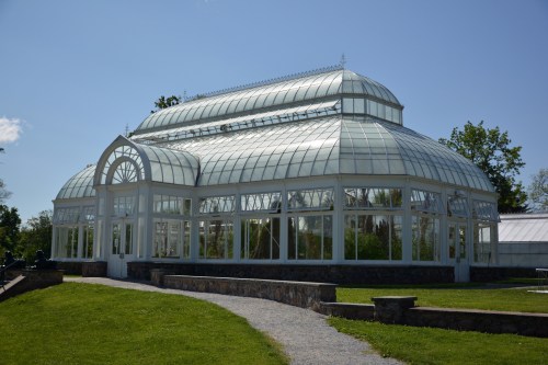 A large glass greenhouse with a curved roof, surrounded by greenery and a stone pathway on a sunny day.