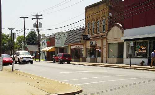 A quiet street scene featuring shops, parked cars, and power lines on a sunny day.