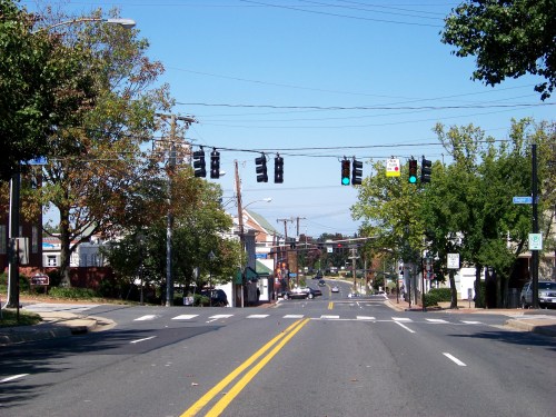 A wide street view with traffic lights, trees, and buildings lining the road under a clear blue sky.