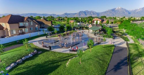 Aerial view of a park with people playing volleyball, surrounded by green grass, trees, and mountains in the background.