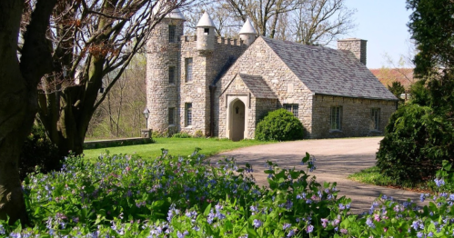 A charming stone cottage with a turret, surrounded by green grass and blooming purple flowers on a sunny day.