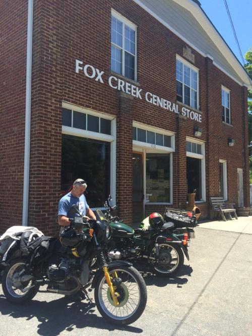 A man adjusts a motorcycle in front of the Fox Creek General Store, a brick building with large windows.