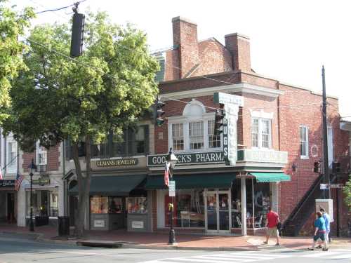 Historic brick building with a pharmacy and jewelry store, featuring large windows and a tree in front.