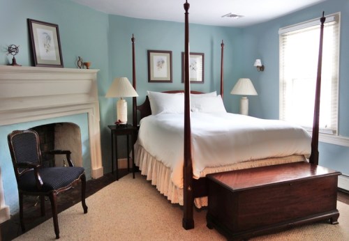 Cozy bedroom featuring a four-poster bed, bedside lamps, a wooden chest, and a chair against a light blue wall.