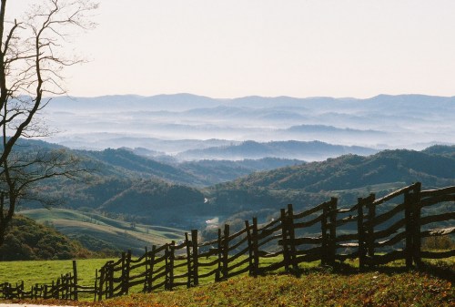 A serene landscape featuring rolling hills, a wooden fence, and misty mountains in the background under a clear sky.