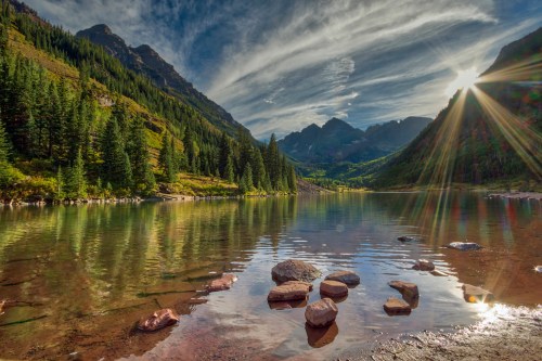 A serene mountain lake surrounded by lush trees, with sunlight reflecting off the water and dramatic clouds above.