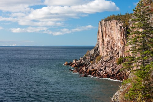 A rocky cliff rises above a calm blue sea under a partly cloudy sky, with trees lining the shore.