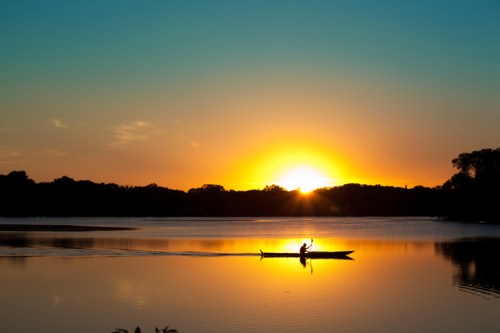A serene sunset over a calm lake, with a lone kayaker paddling towards the horizon.