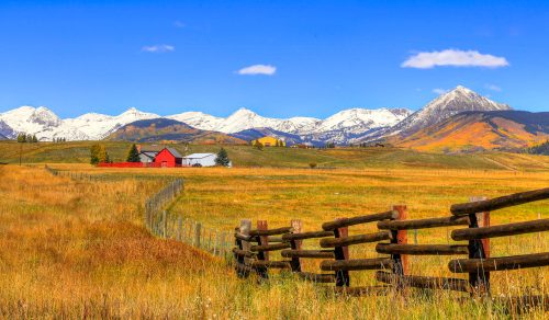 A scenic landscape featuring a red barn, a wooden fence, and snow-capped mountains under a clear blue sky.