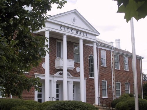 A brick city hall building with white columns and a sign reading "CITY HALL," surrounded by green shrubs and trees.