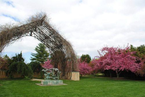 A lush garden featuring a vine-covered arch and blooming pink trees, with a sculpture in the foreground.