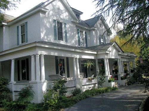A large, white Victorian-style house with a wraparound porch and lush greenery surrounding it.