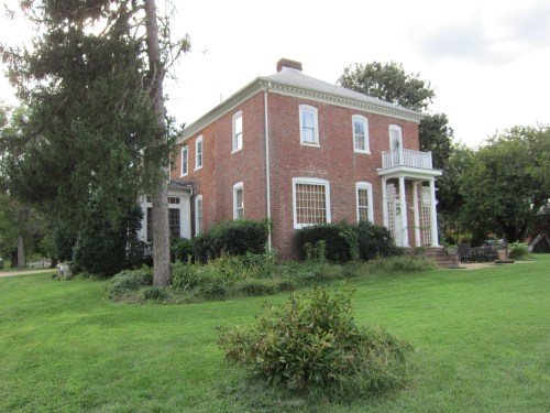 A two-story brick house surrounded by greenery and trees, featuring a front porch and large windows.