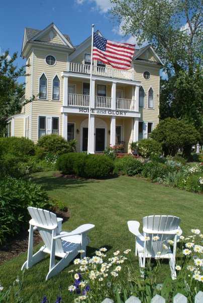A charming yellow house with an American flag, surrounded by lush gardens and two white Adirondack chairs in the foreground.