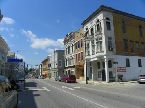 A street view featuring historic buildings, blue skies, and parked cars in a small town.