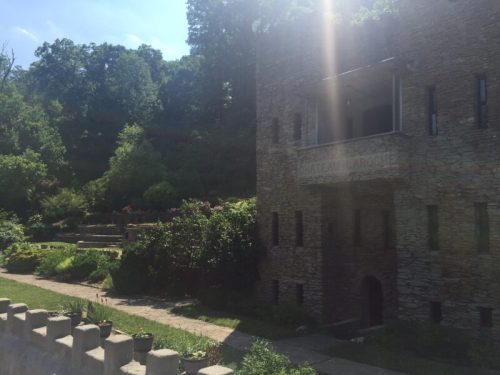 A stone building with a sign, surrounded by lush greenery and gardens under bright sunlight.