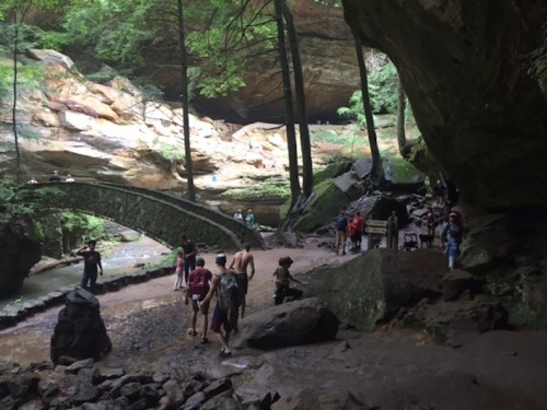 A scenic forest area with a stone bridge, people walking along a stream, and lush greenery surrounding the rocky landscape.