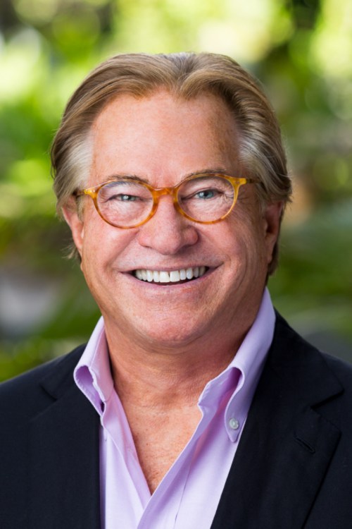 Smiling man with glasses, wearing a black blazer over a light purple shirt, against a blurred green background.