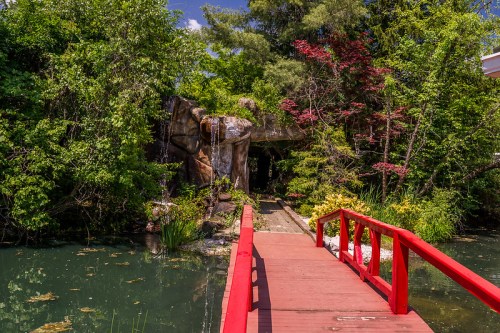 A red wooden bridge leads through lush greenery to a rocky archway over a serene pond.