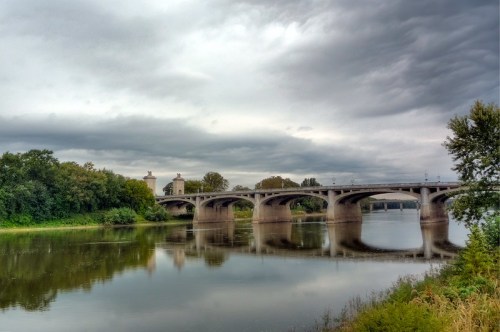 A stone bridge arches over a river, reflecting in the water under a cloudy sky, surrounded by greenery.