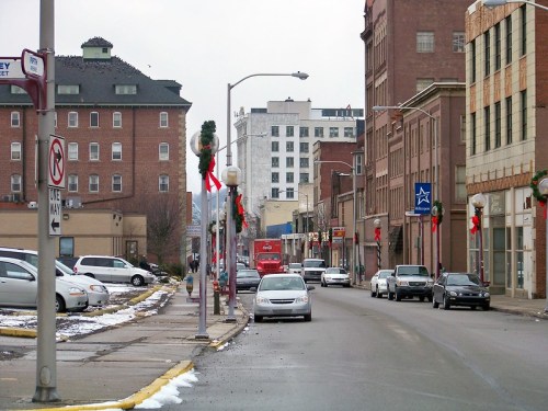 A city street decorated for the holidays, featuring cars parked along the road and buildings in the background.
