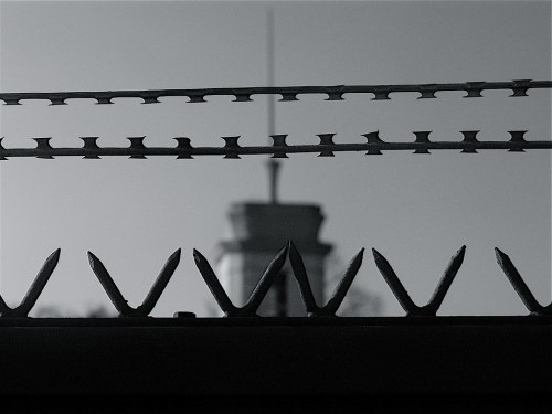 Black and white image of barbed wire in the foreground with a blurred tower in the background.