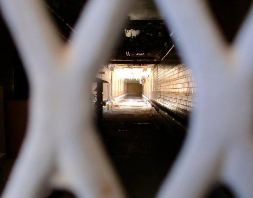 A dimly lit corridor viewed through a white metal grate, with light at the far end illuminating the space.