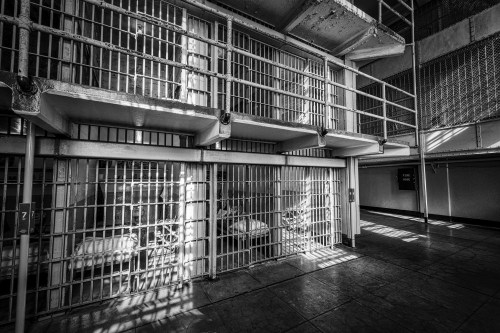 Black and white image of a prison interior, featuring barred cells and shadows cast by sunlight.