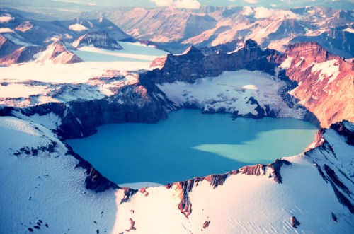 Aerial view of a volcanic crater lake surrounded by snow-capped mountains and glaciers under a clear sky.